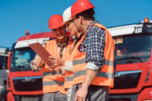 Three workers in orange high-visibility jackets examine a clipboard standing in front of red trucks.